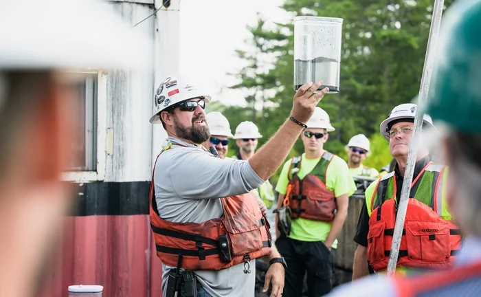 A Brennan crew member in a hard hat and life vest shows a water container to a group during an environmental project.