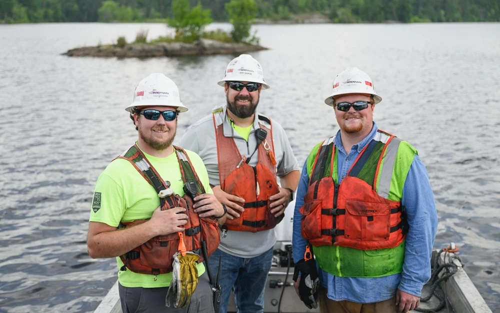 A Brennan crew in safety gear and life vests performs environmental remediation work from a boat on a lake.