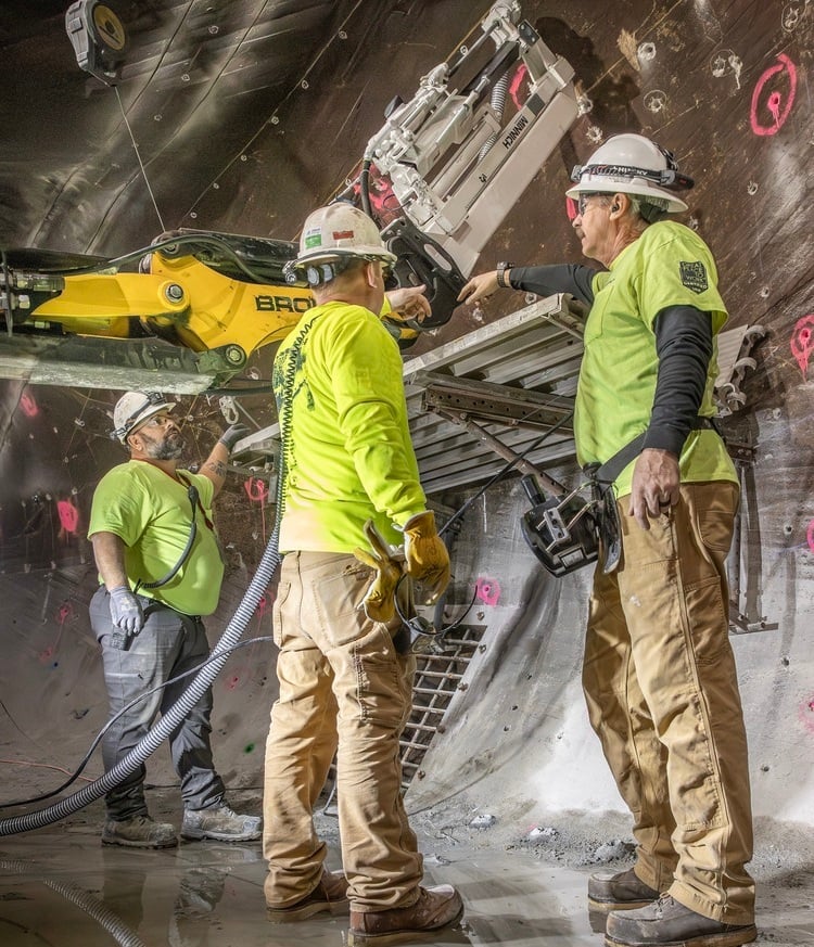 A Brennan crew performs concrete rehabilitation using a rock drill on yellow machinery inside a curved tunnel.