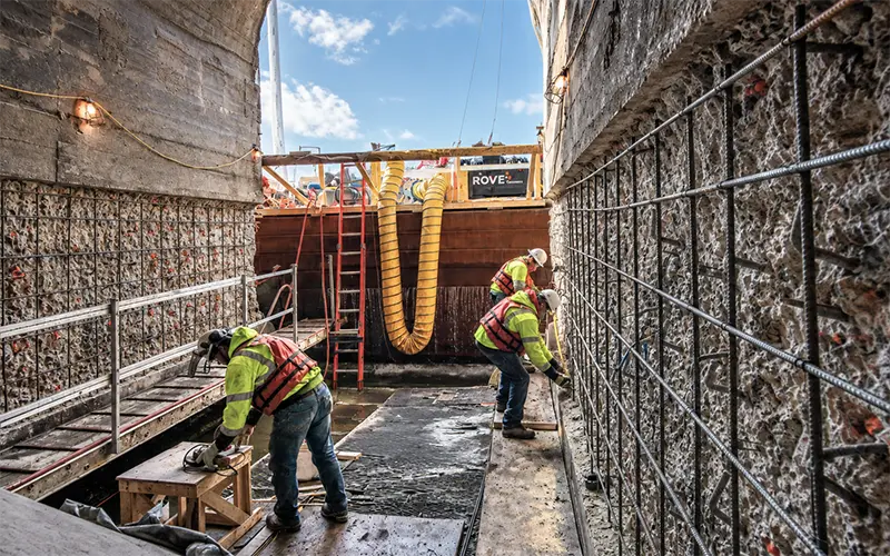 Brennan crew members in safety gear install reinforcing steel against concrete walls in a narrow excavation.