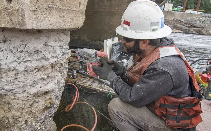 A Brennan worker in safety gear uses a handheld hammer drill to bore into a concrete bridge pier above the water.