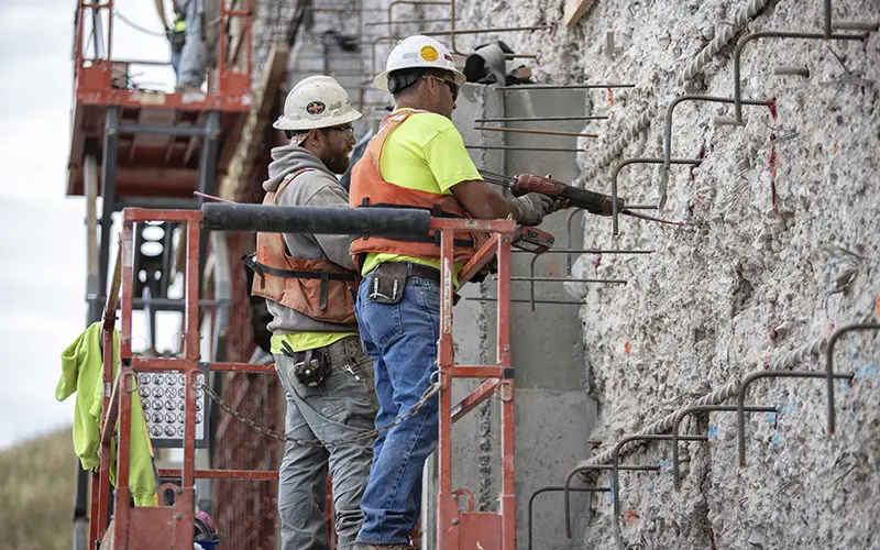 Brennan crew members in safety gear use a power tool to drill into a concrete retaining wall from a lift.